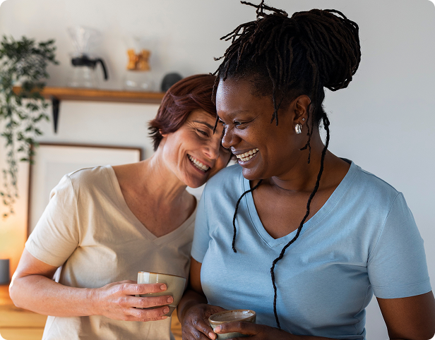Two women laughing joyfully together while holding coffee mugs indoors.