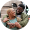 Smiling Black man warmly hugging a woman with short blonde hair from behind in a circular frame.