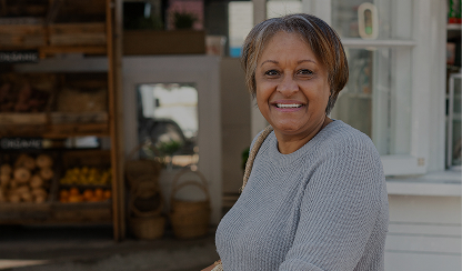 Smiling older woman carrying a shoulder bag while standing outside a fresh produce market.